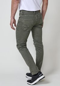 Homme portant un jean gris slim et des baskets noires, debout avec une jambe légèrement pliée devant un fond uni.