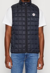 Marinblå pufferväst med quiltat mönster, hög krage, dragkedjestängning och logotypdetalj på vänster bröst, bärs över en vit t-shirt.