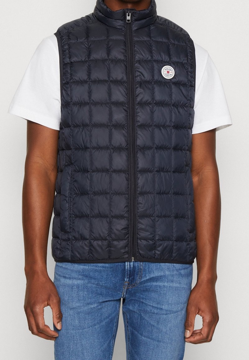 Marinblå pufferväst med quiltat mönster, hög krage, dragkedjestängning och logotypdetalj på vänster bröst, bärs över en vit t-shirt.