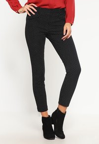 Leggings noirs ajustés avec des pois blancs, présentant une texture lisse et une taille mi-haute, associés à des bottes noires à chevilles.