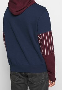 Mörkblå hoodie med en vinröd rand på ärmen, med kapuschong och ribbad mudd, tillverkad av mjukt bomullsmaterial.