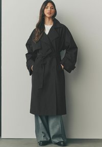 Svart trenchcoat med bälte i midjan, breda slag och långa ärmar. Bärs över grå vida byxor, i kombination med svarta skor.