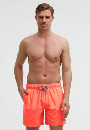 Oberkörperfreier Mann mit kurzen braunen Haaren, der leuchtend orange Badeshorts mit einem schwarz-weißen Kordelzug trägt und vor einem schlichten Hintergrund steht.