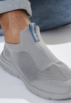 Light gray slip-on knit sneaker with flexible sole, blue and white pull tab, worn with cuffed light blue pants.