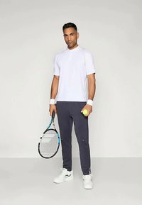 Bijela majica kratkih rukava za sport, tamno sivi hlače, drži žuti teniski lopticu i reketa s plavim akcentima, stoji na neutralnoj pozadini.