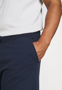 Marinblå joggerbyxor med mjuk textur, som har midjeband med dragsko och sidofickor. Dekorativ sömnad framhäver designen.