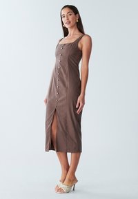 Braunes Leinen-Midikleid mit quadratischem Ausschnitt, vorderer Knopfleiste und seitlichem Schlitz. Modell trägt offene, geflochtene High Heels.