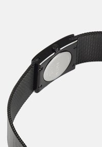 Orologio con cinturino in rete nera che presenta una cassa rettangolare, una lunetta lucida e un retro argentato. Il cinturino ha un motivo tessuto e testurizzato.