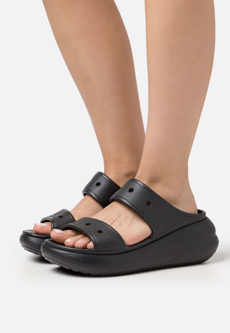 Pieds portant des sandales compensées noires à enfiler avec deux larges brides et de petits trous de ventilation sur fond blanc.
