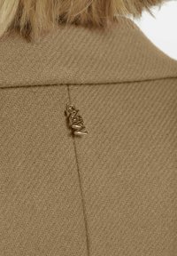 Bruine gestructureerde stof met een subtiel visgraatpatroon. Kleine metalen logo-accent op de achterkant nabij de naad. Strak, verzorgd ontwerp.