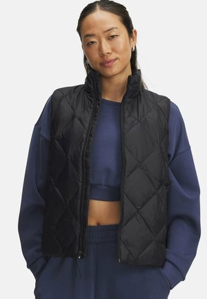 Jonge vrouw die een zwart gewatteerd vest draagt over een blauw cropped sweatshirt en bijpassende broek, staand met handen in de zakken.