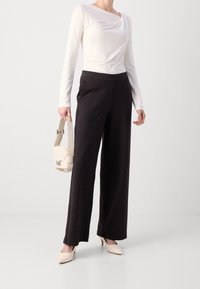 Witte langemouwen top met gedrapeerde halslijn, gecombineerd met hoge zwarte broek met pinstripe en crèmekleurige hakken, met een kleine tas in de hand.