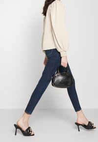 Černé lakované heeled mules s zlatým akcentem, spárované s tmavě modrými skinny džínami a malou černou texturovanou kabelkou.