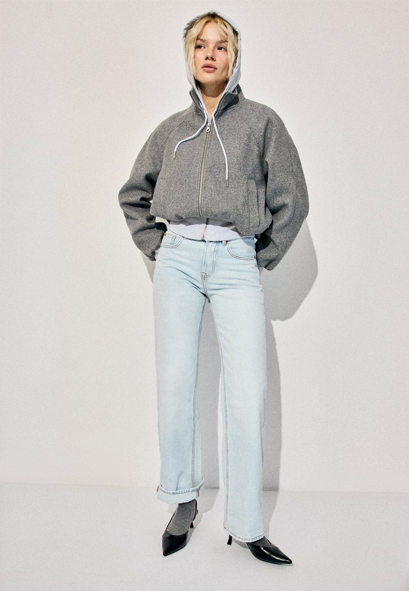 Vaaleahiuksinen nainen seisoo käsine taskuissa, päällään harmaa lyhyt vetoketjullinen huppari, vaaleansiniset suorat farkut, harmaat sukat ja mustat teräväkärkiset korkokengät.