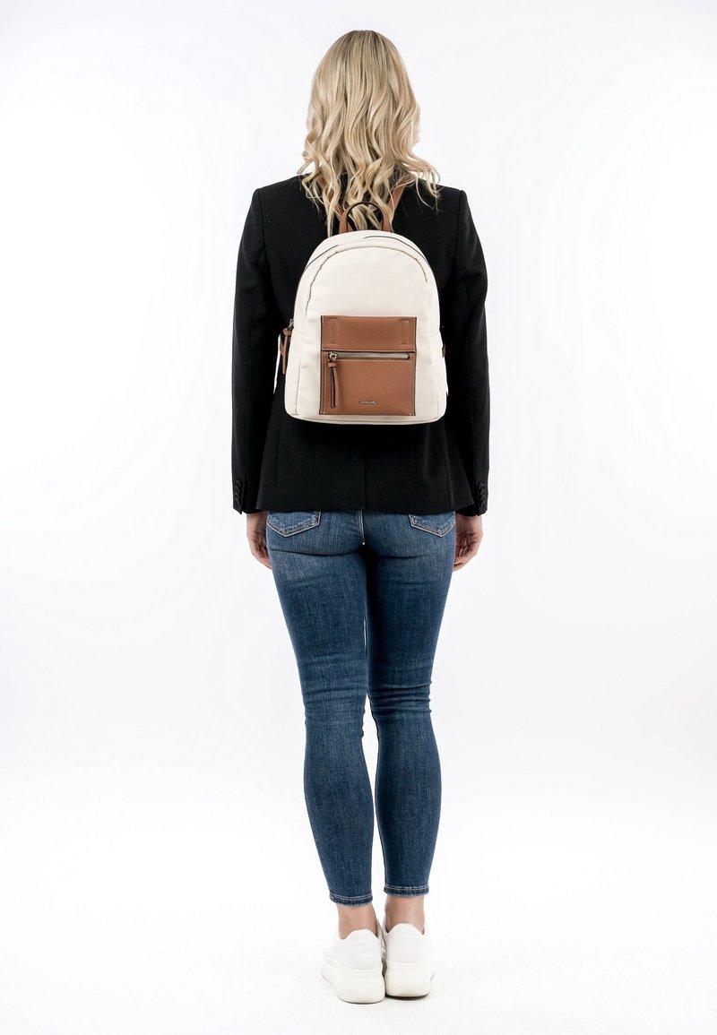 Femme aux cheveux blonds mi-longs portant une veste noire, un jean bleu, des chaussures blanches et un sac à dos beige avec une poche avant marron, vue de dos.