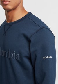 Felpa blu navy realizzata in tessuto morbido, presenta lettere in rilievo e dettagli del logo sul petto e sulla manica. Collo e polsini a coste.