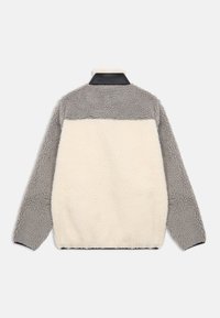 Fleece striukė su pilkais rankovėmis ir viršutine nugaros dalimi, kreminės spalvos apatine nugaros dalimi ir juodu apykakle prieš baltą foną.