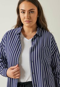Camisa de botones a rayas azul y blanca, con un bolsillo en el pecho, mangas largas y un corte relajado. Llevada sobre una camiseta blanca.
