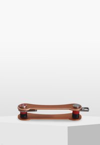 L'outil présente un corps incurvé de couleur bronze avec un accent en métal argenté, des détails en caoutchouc rouge et noir, positionné sur une surface blanche.