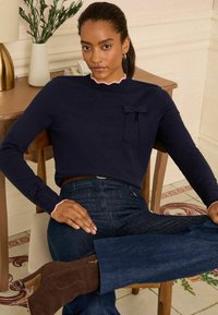 Námořnický dlouhý rukáv top s vroubkovaným výstřihem, přední mašlí a růžovými akcenty; nošený s tmavými denimovými džíny a hnědými kotníkovými botami.