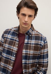 Camicia di flanella a quadri con motivi blu, marrone e bianco, colletto con bottoni e tasca frontale. Indossata sopra una maglietta color granata.