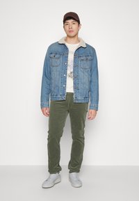 Denim jaka ar kažokādas apkakli, gaiši zila krāsa; bežs grafiskais t-krekls; zaļas korduroja bikses; pelēki sporta apavi; brūna kepka.