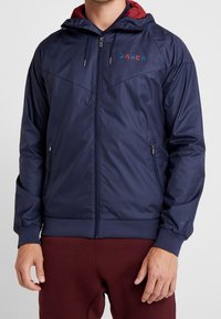 Navyblauwe lichtgewicht jas met een rode gevoerde capuchon, ritssluiting en elastische manchetten. Voorzien van geborduurd logo op de borst.