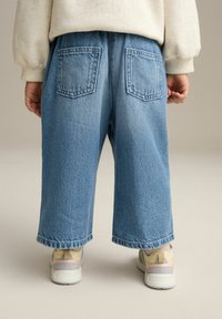 Pantalons larges en denim bleu avec deux poches arrière, featuring un ourlet brut et une texture délavée. Associés à des baskets pastel.