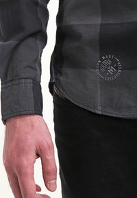 Schwarz-grau kariertes Hemd mit Knopfkragen und einer gestickten Logodetailnaht nahe dem Saum. Der Stoff wirkt glatt und fein.