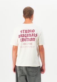 Jaunas asmuo vilkintis off-white spalvos marškinėlius su dideliu raudonu užrašu "STUDIO ORIGINALS EDITION" ir mažesniu užrašu žemiau, derinant su pilkomis kelnėmis.