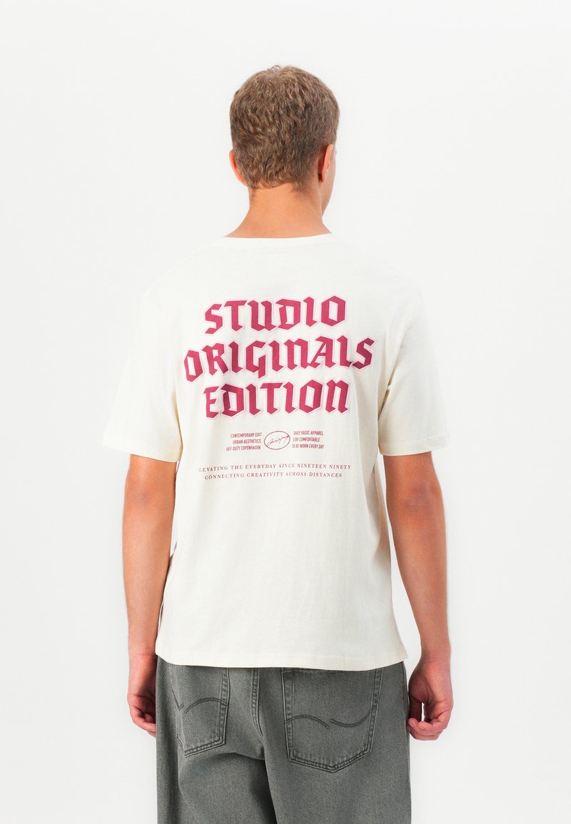 Jaunas asmuo vilkintis off-white spalvos marškinėlius su dideliu raudonu užrašu "STUDIO ORIGINALS EDITION" ir mažesniu užrašu žemiau, derinant su pilkomis kelnėmis.