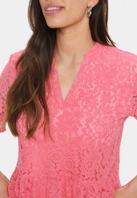 Rožinė nėriniuota palaidinė su trumpomis rankovėmis ir V formos iškirpimu; turi gėlių raštus ir tekstūruotą paviršių. Diskretus volanas apatinėje dalyje.