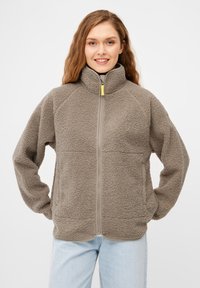 Flauschige beiger Fleecejacke mit hohem Kragen, frontalem Reißverschluss und grauen Akzenten. Verfügt über Seitentaschen und eine strukturierte Oberfläche.