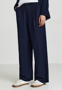 Navy-blaue weite Hosen mit vertikalen Streifen, gefaltetem Vorderteil, Seitentaschen und elastischem Bund, kombiniert mit weißen und grauen Sneakers.
