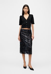 Μαύρο crop top με βολάν στα μανίκια και κουμπιά μπροστά, σε συνδυασμό με μαύρη midi φούστα με παγιέτες και μυτερά μαύρα τακούνια.