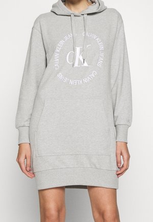 Kobieta nosząca szarą sukienkę z kapturem Calvin Klein z przednią kieszenią i logo w białym kole na piersi.