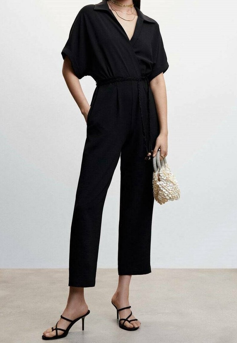 Femme portant une combinaison noire ceinturée à manches courtes, des talons noirs à brides et tenant un petit sac à main crème texturé.