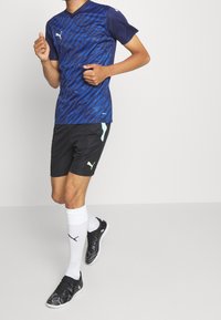 Blåmönstrad sporttröja med kort ärm, i kombination med svarta shorts med ljusblå accent, vita strumpor och svarta träningsskor.