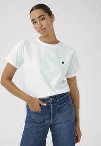Camiseta blanca de algodón con mangas cortas enrolladas, que presenta un pequeño diseño de corazón negro en el lado izquierdo del pecho. Llevada con vaqueros azules de cintura alta.