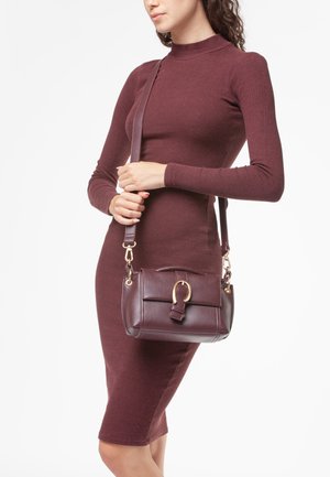 Donna che indossa un vestito bordeaux a maniche lunghe aderente, mentre tiene una borsa di pelle coordinata con una fibbia dorata e una tracolla.