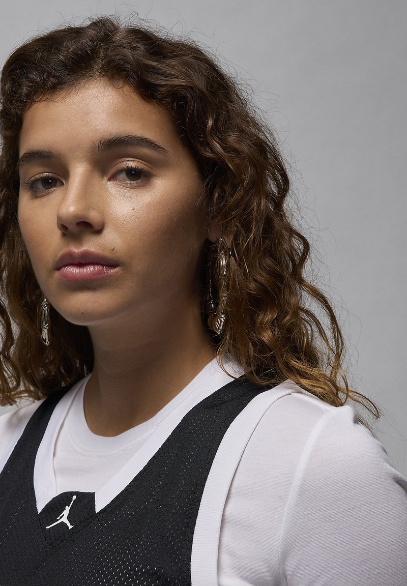 Schwarzes Mesh-Trikot mit weißen Akzenten über einem weißen Shirt, mit einem kleinen Logo. Das Model hat lockige Haare und trägt hängende Ohrringe.