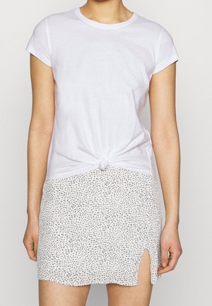 Woman wearing a white short-sleeve t-shirt tied at the waist and a white skirt with small black spots and a front slit on the right side.