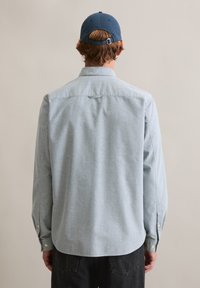 Jasnoniebieska koszula z długim rękawem wykonana z miękkiej tkaniny, o prostym kroju, z mankietami na guziki i detalem na karku. Noszona z granatową czapką.