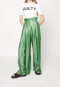 Pantalones de satén verdes de pierna ancha con cintura alta y costuras laterales sutiles. Combinados con una camiseta blanca con texto negro. Calzado negro.