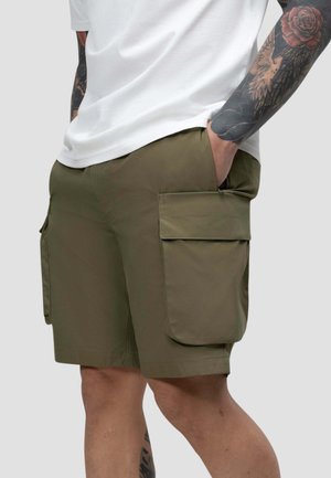 Mann trägt olivgrüne Cargo-Shorts und ein weißes Hemd, hat tätowierte Arme, Hände in den Taschen, vor schlichtem Hintergrund.