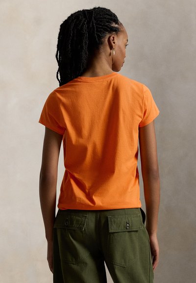 Κοντομάνικο πορτοκαλί βαμβακερό μπλουζάκι με στρογγυλή λαιμόκοψη, εμφανιζόμενο από πίσω, σε συνδυασμό με πράσινο παντελόνι καμπαρντίνα.