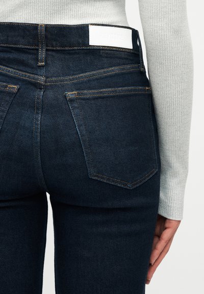 Σκούρο μπλε τζιν με ορατές πίσω τσέπες, αντίθετη ραφή και λευκή ετικέτα μάρκας στη μέση, φορεμένο με ανοιχτό γκρι ριμπ τοπ.