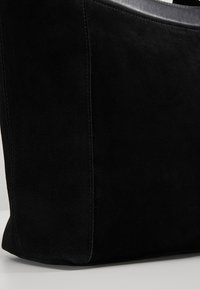 Sac à main en daim noir avec bord supérieur bordé de cuir, forme rectangulaire et coutures apparentes sur les côtés.