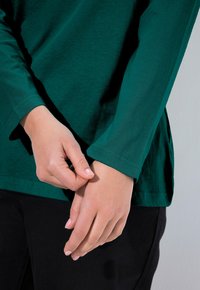 Zielona koszula z długim rękawem o gładkiej fakturze. Rękawy są dopasowane, a dolny brzeg jest lekko zaokrąglony, w zestawieniu z czarnymi spodniami.