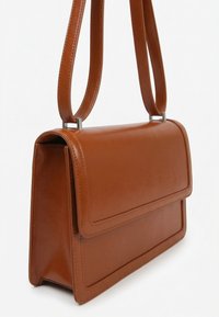 Sac à épaule rectangulaire en cuir marron avec fermeture à rabat et double bandoulière suspendu sur un fond blanc uni.